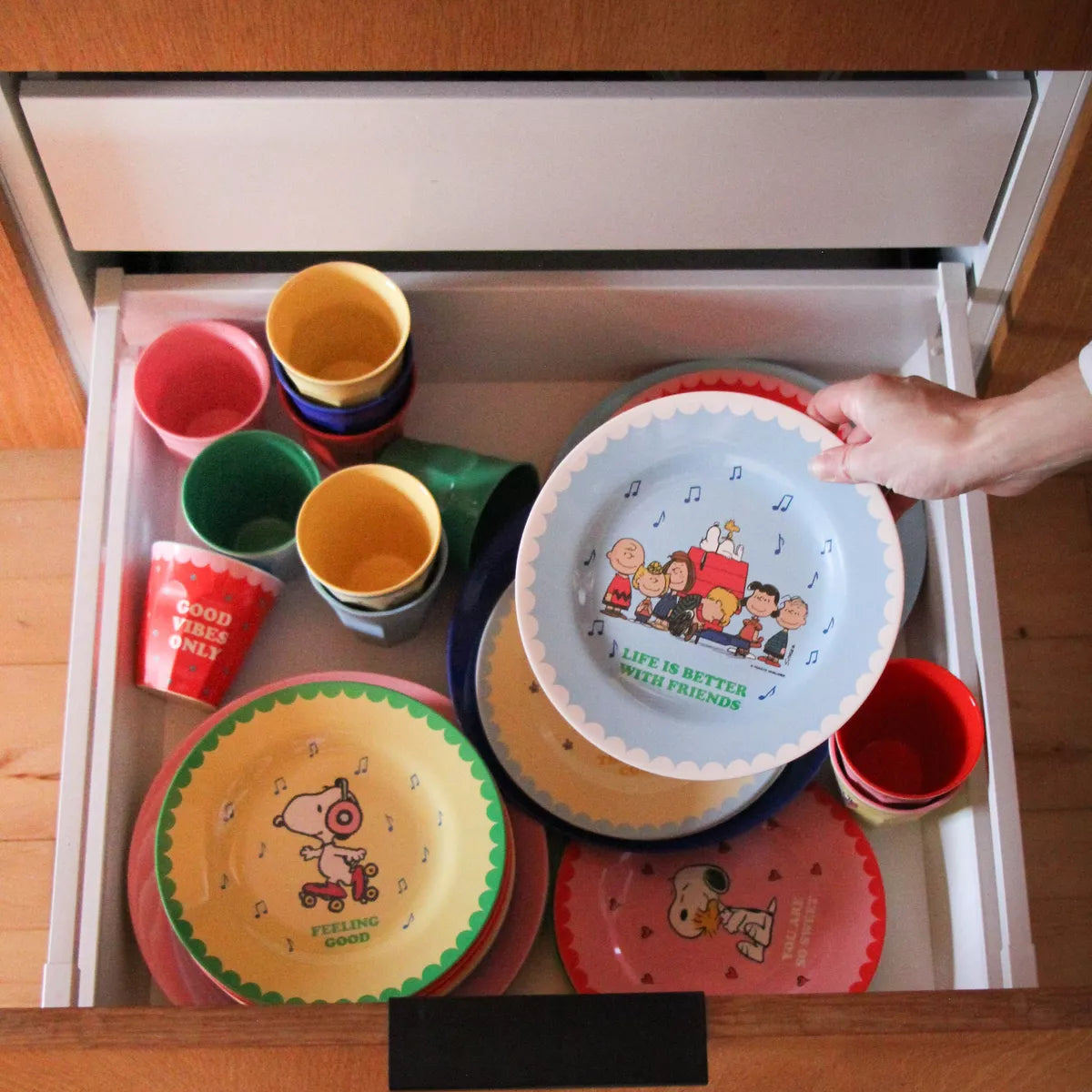 A hand holding a light blue Snoopy-themed plate inside an open wooden drawer filled with colorful children's plates and cups.