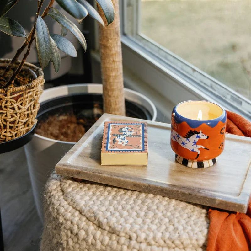 Candle with decorative packaging on a wooden tray next to potted plants