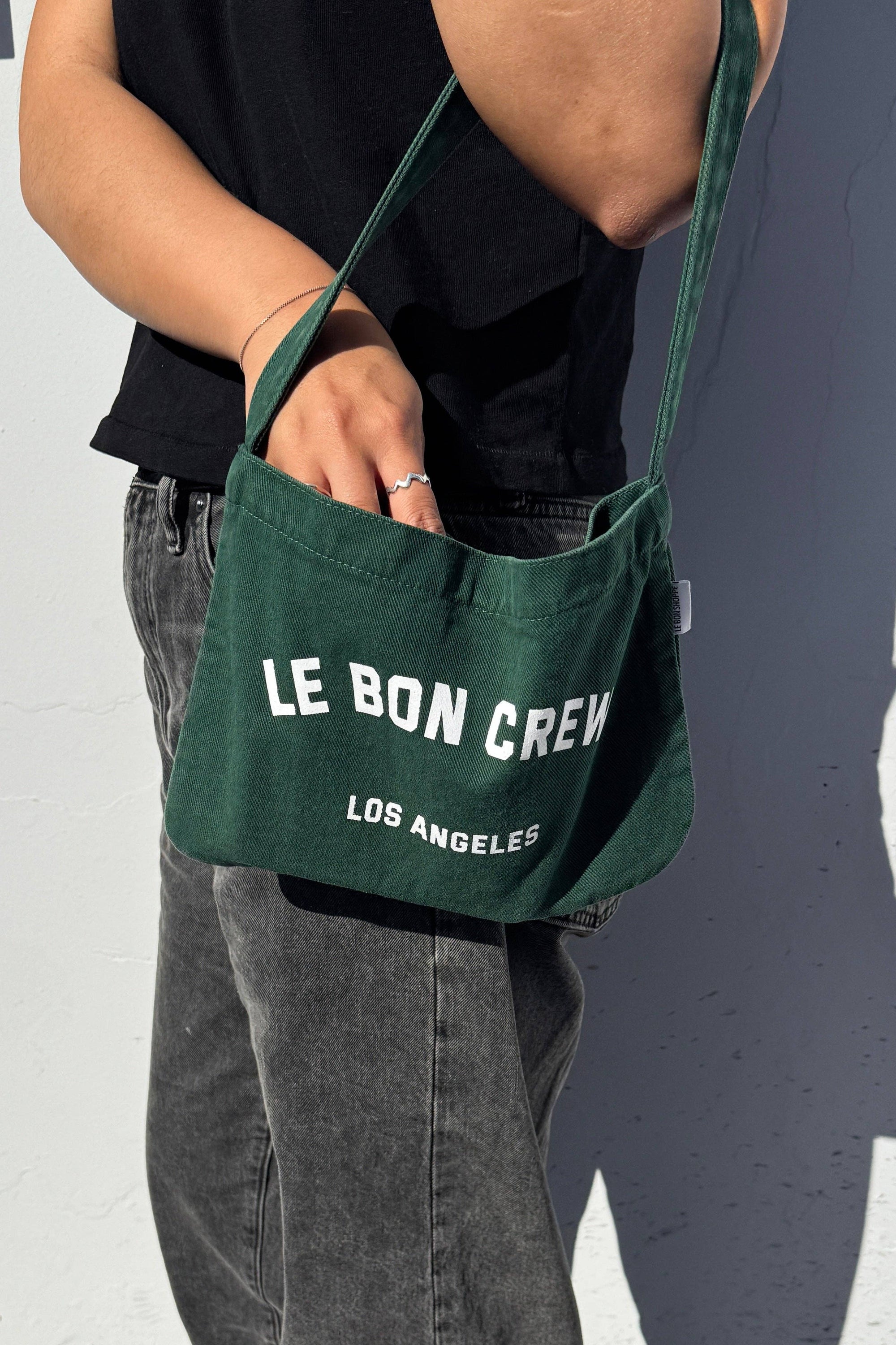 Person holding a green bag with 'Le Bon Crew Los Angeles' text against a plain background